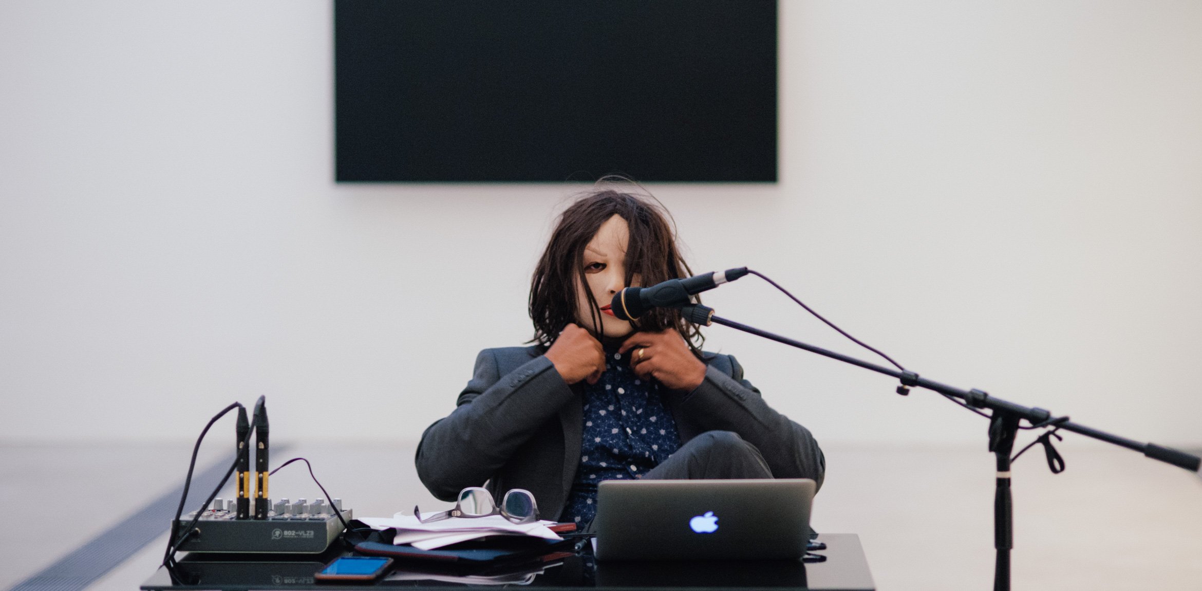 Ronaldo Wilson sits in a mask behind a desk with a microphone and laptop, in front of Ellsworth Kelly's "Blue Black."