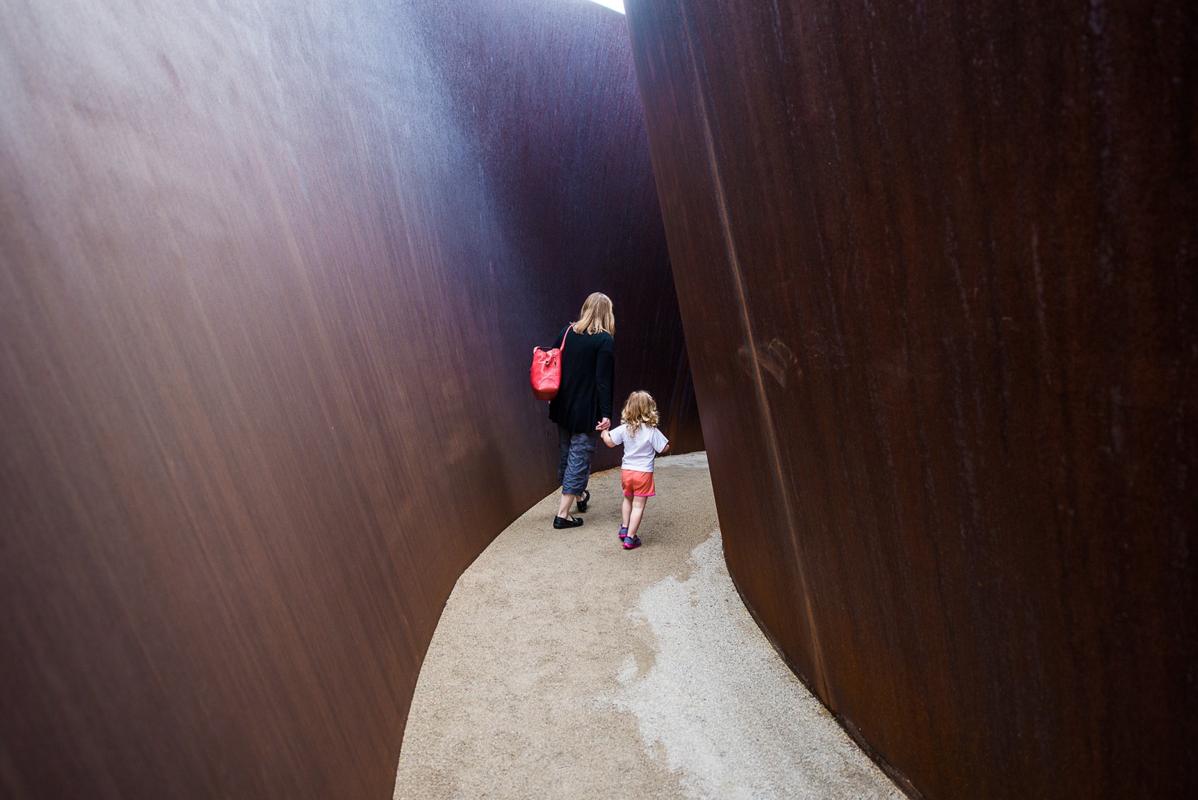 A parent and child hold hands and walk inside Richard Serra's "Joe."