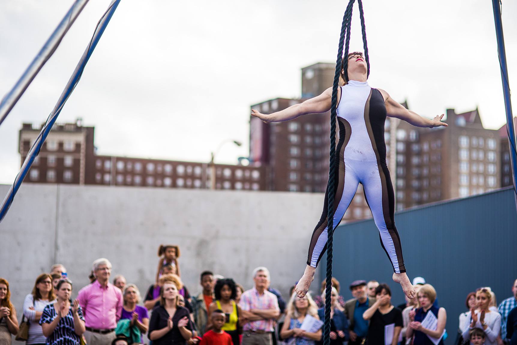 In the Courtyard, an acrobat floats in the air for a large audience with their arms and legs outstretched, the nape of their neck resting in a blue rope.