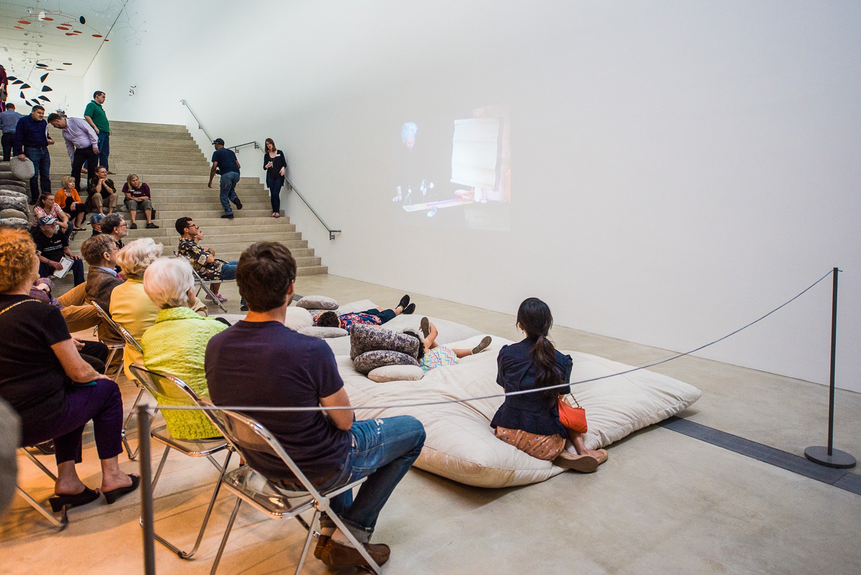 An audience, some seated in chairs and some lying on a large cushion, watches a film projected on the wall of the Lower-Main Gallery.
