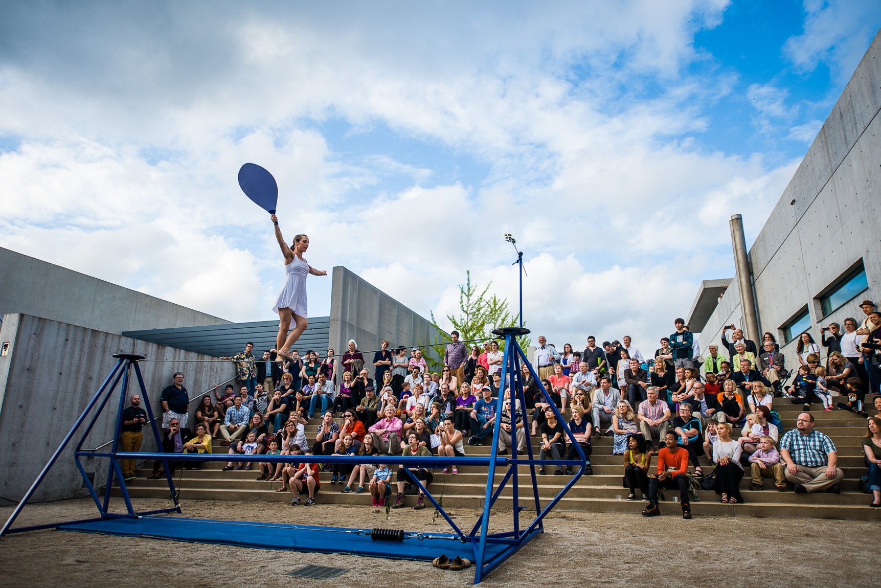 An acrobat holding a blue balancing fan walks on a tightrope in the Courtyard for a large audience gathered on the stairs.