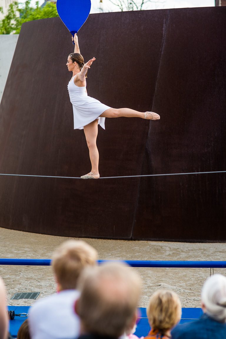 An acrobat holding a blue balancing fan arabesques on a tightrope in front of Serra's "Joe" for an audience.