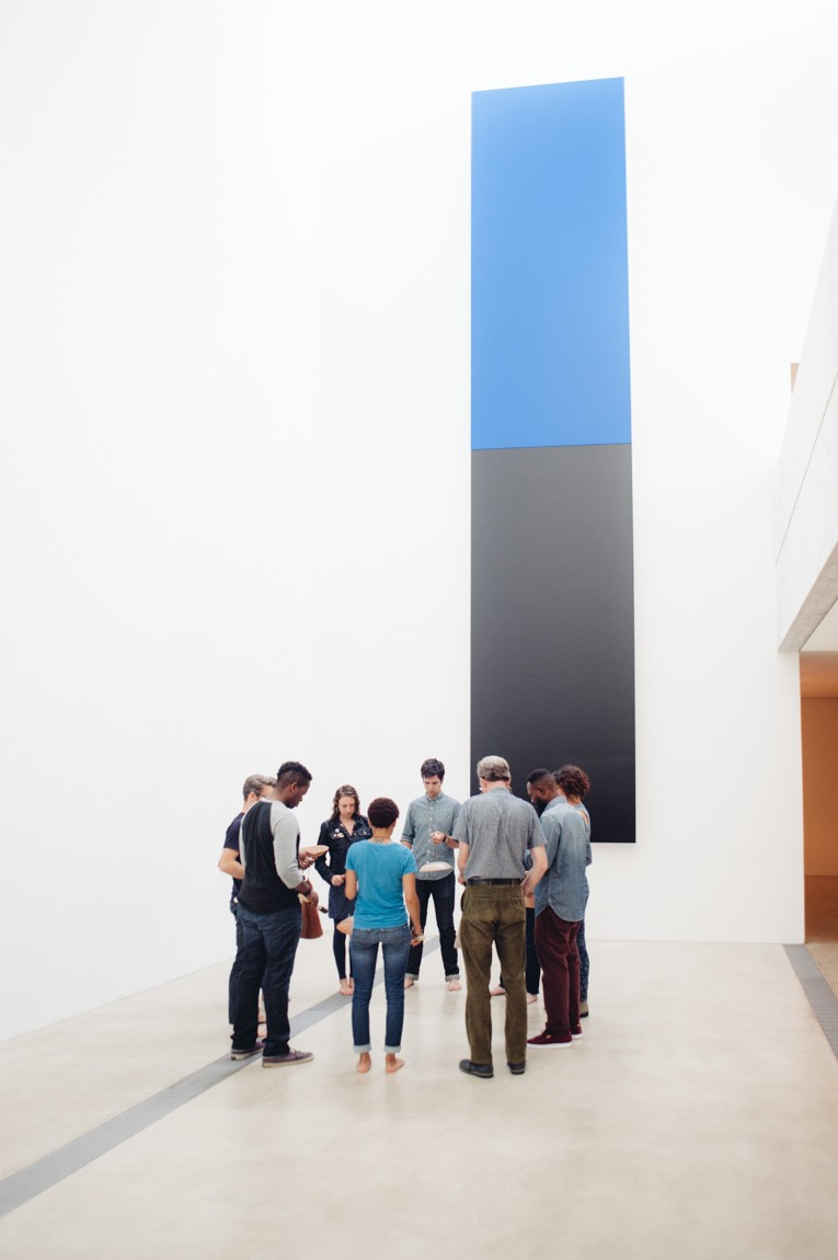 Participants hold clay instruments and drum on them with wooden mallets in a circle of people facing inwards, in front of Kelly's "Blue Black."