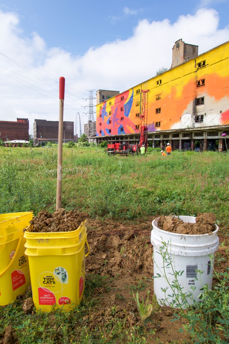 Three buckets filled with Mississippi River clay. In the background there's a colorful building and the Arch.