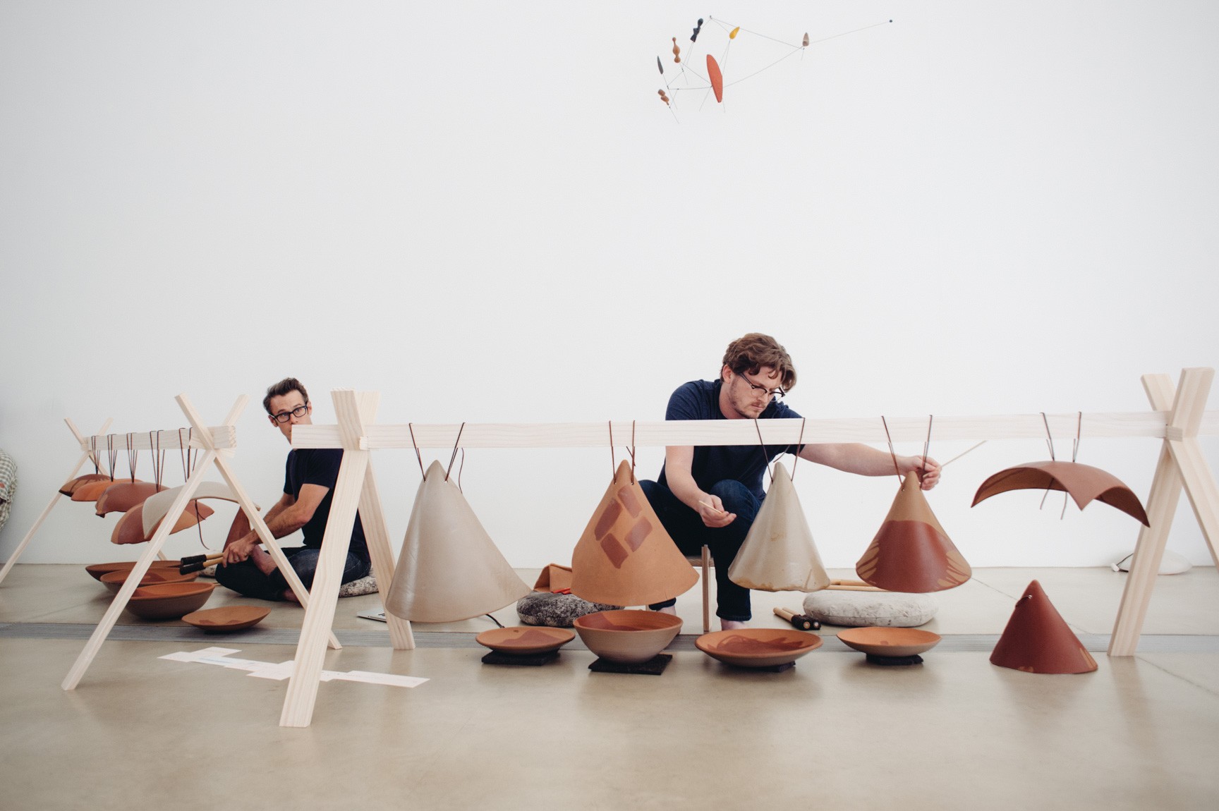 Chris Kallmyer and an assistant kneel behind a white beam holding hanging clay instruments, using percussive sticks to tap the clay.