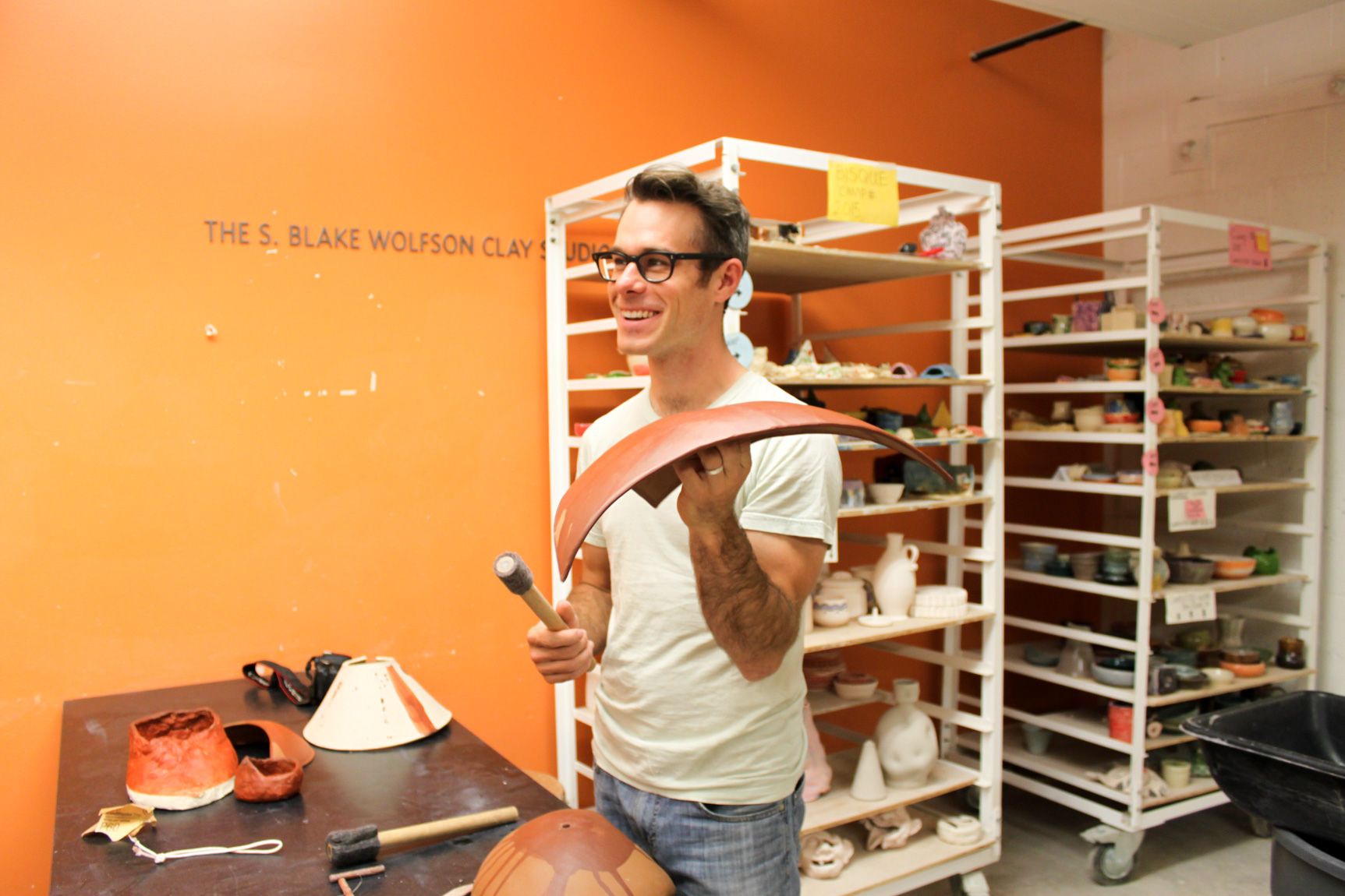 Kallmyer in his studio, smiling and holding a long, rectangular clay instrument in his left hand, and a felted mallet in his right.