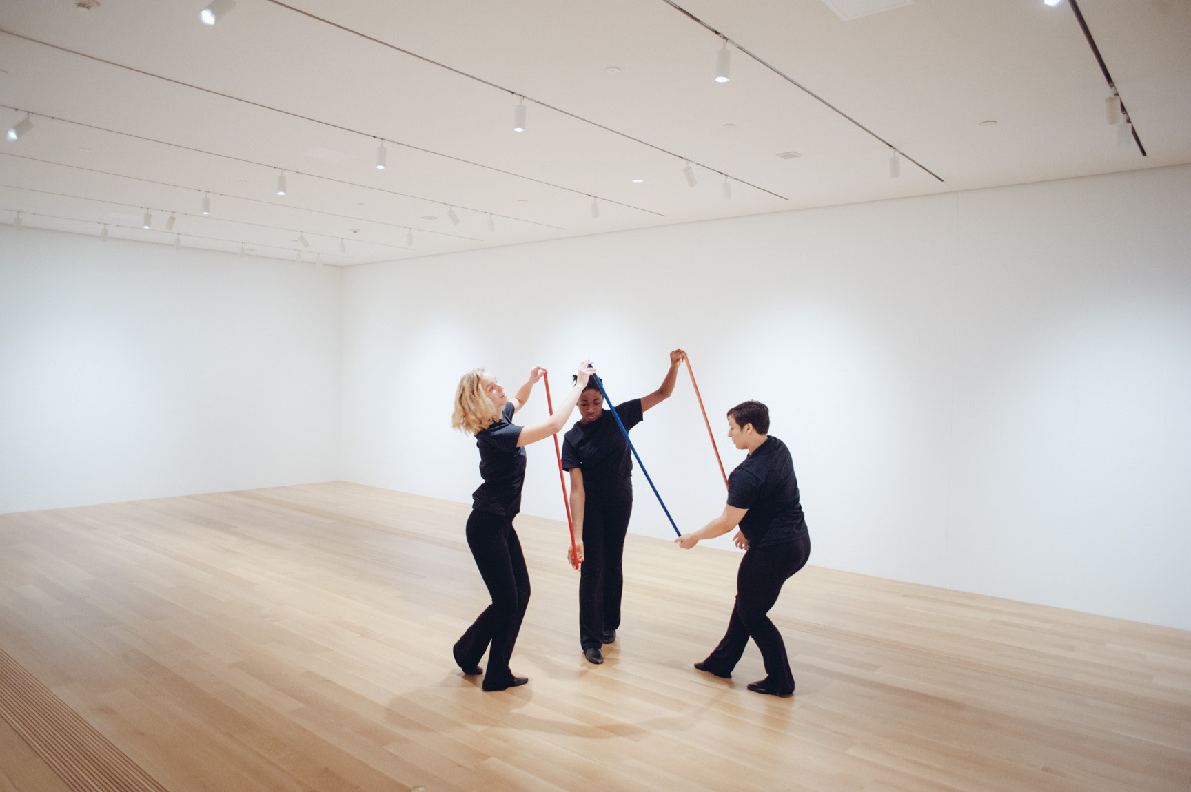 Three students perform in the Lower-East Gallery holding three red, white, and blue sticks interlocking the performers.