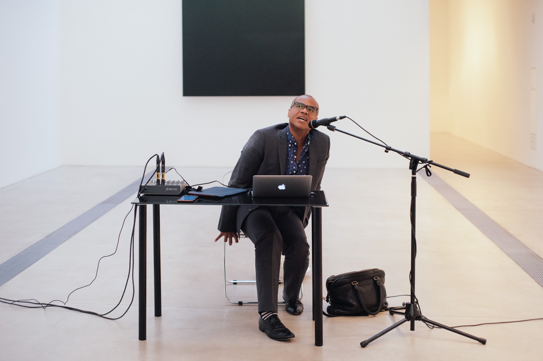 Ronaldo Wilson sits behind a desk with a microphone, with Ellsworth Kelly's "Blue Black" in the background.