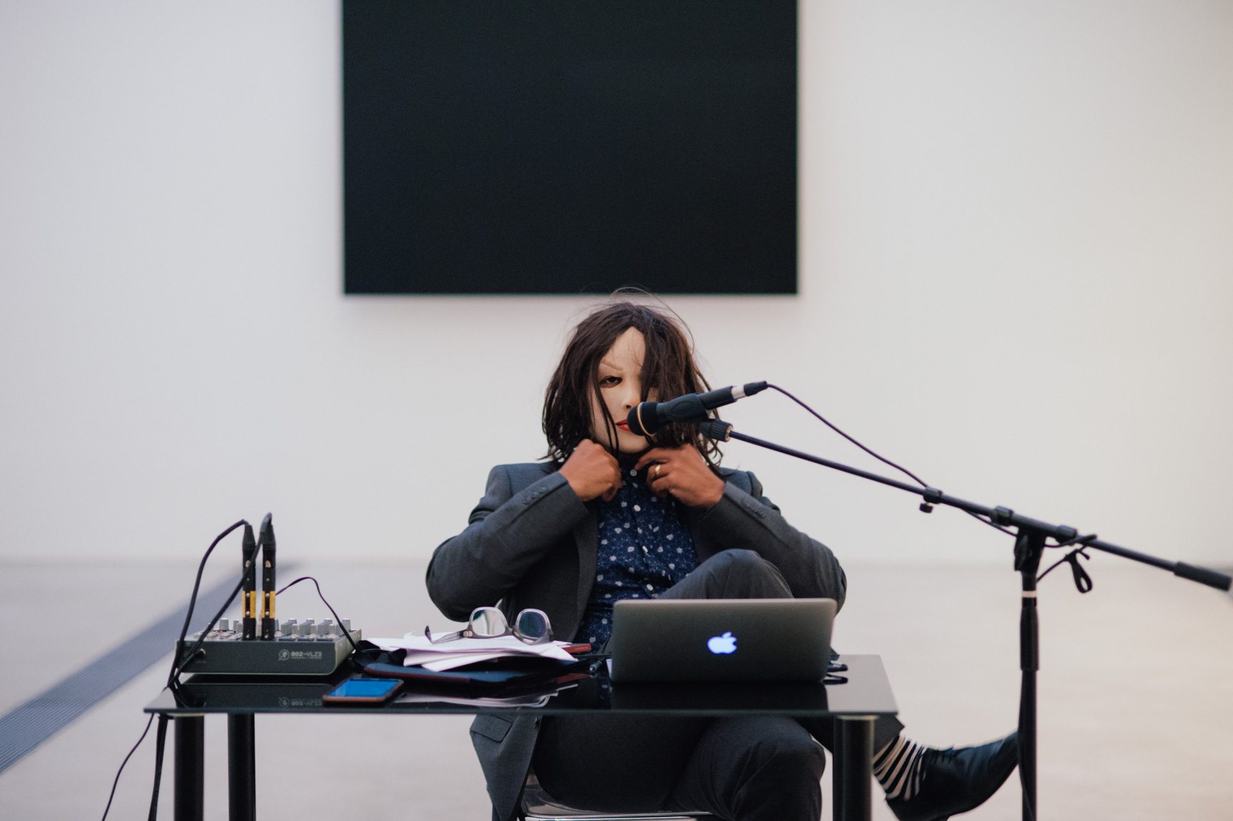 Ronaldo Wilson sits in a mask behind a desk with a microphone. Behind them is Ellsworth Kelly's "Blue Black."