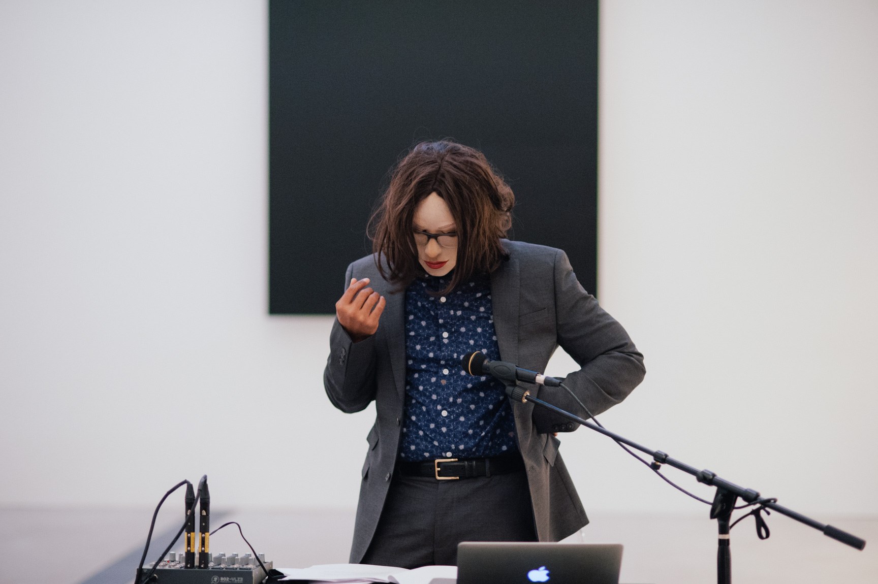 Ronaldo Wilson stands in costume behind a desk looking at their notes and speaks into a microphone. Behind them is Ellsworth Kelly's "Blue Black."