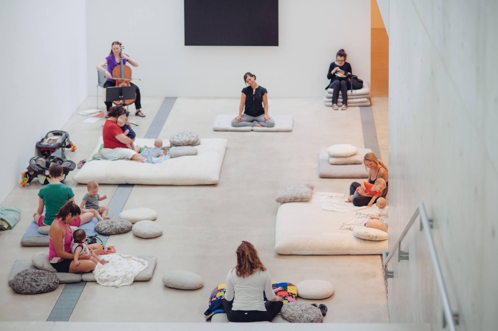 Visitors and their infants participate in an exercise on cushions in the Lower-Main Gallery, while a cellist plays in the corner.