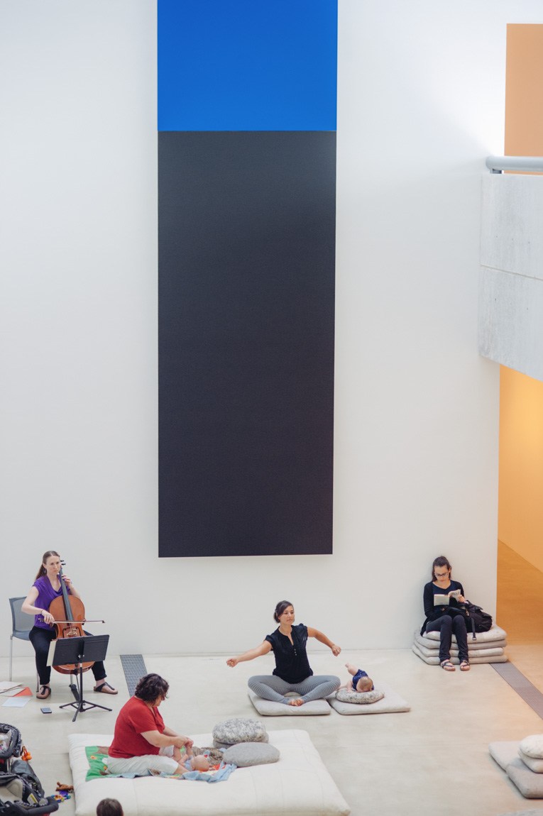 Visitors and their infants are led by Stacy Broussard in a yoga exercise, on cushions in the Lower-Main Gallery in front of "Blue Black" by Ellsworth Kelly, while a cellist plays in the corner.