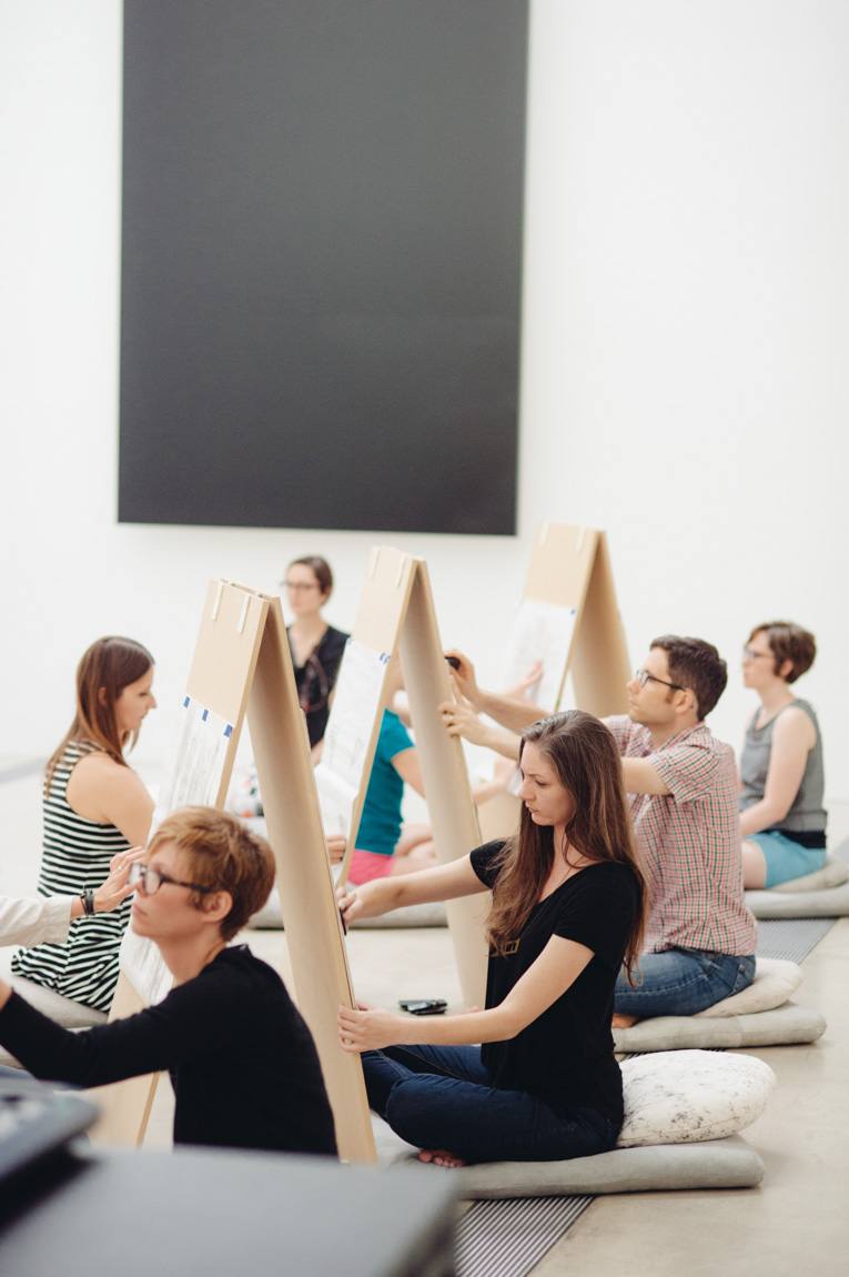 Participants sit on cushions and draw on either sides of a triangular easel in the Lower-Main Gallery.