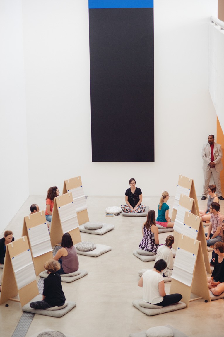 A view of Angela Malchionno leading participants in meditation in front of "Blue Black" by Ellsworth Kelly. Participants sit in front of easels.