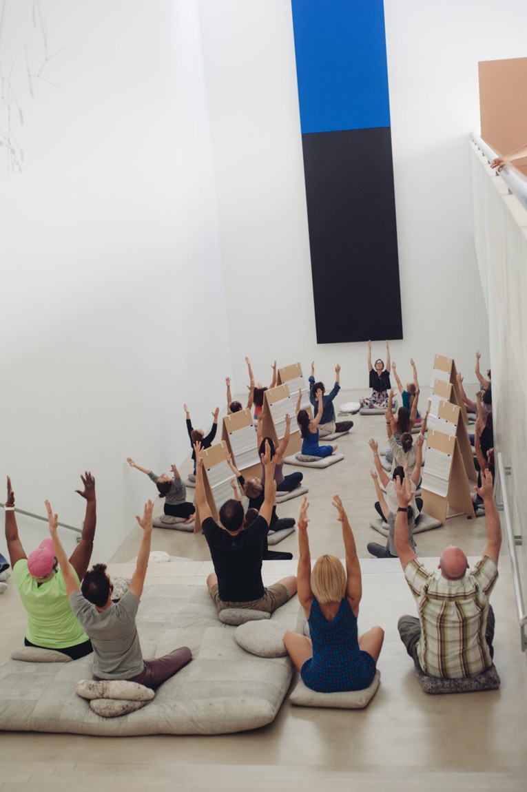 Angela Malchionno leads a large group in meditation in front of Kelly's "Blue Black." The whole group is cross-legged and raising their arms straight above their heads.
