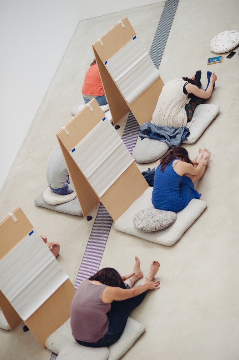 Participants meditate on cushions in front of easels with their legs out and head stretched to touch their knees in the Lower-Main Gallery.