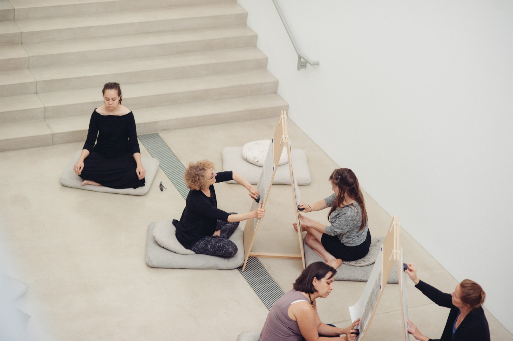 Participants sit on cushions and draw on either sides of a triangular easel in the Lower-Main Gallery. Another meditates cross-legged on a cushion at the foot of the Main Staircase.