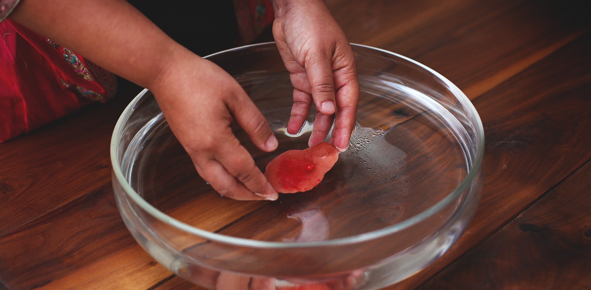 A closeup of Bhanu Kapil's hands placing a piece of clay into a clear bowl of water.