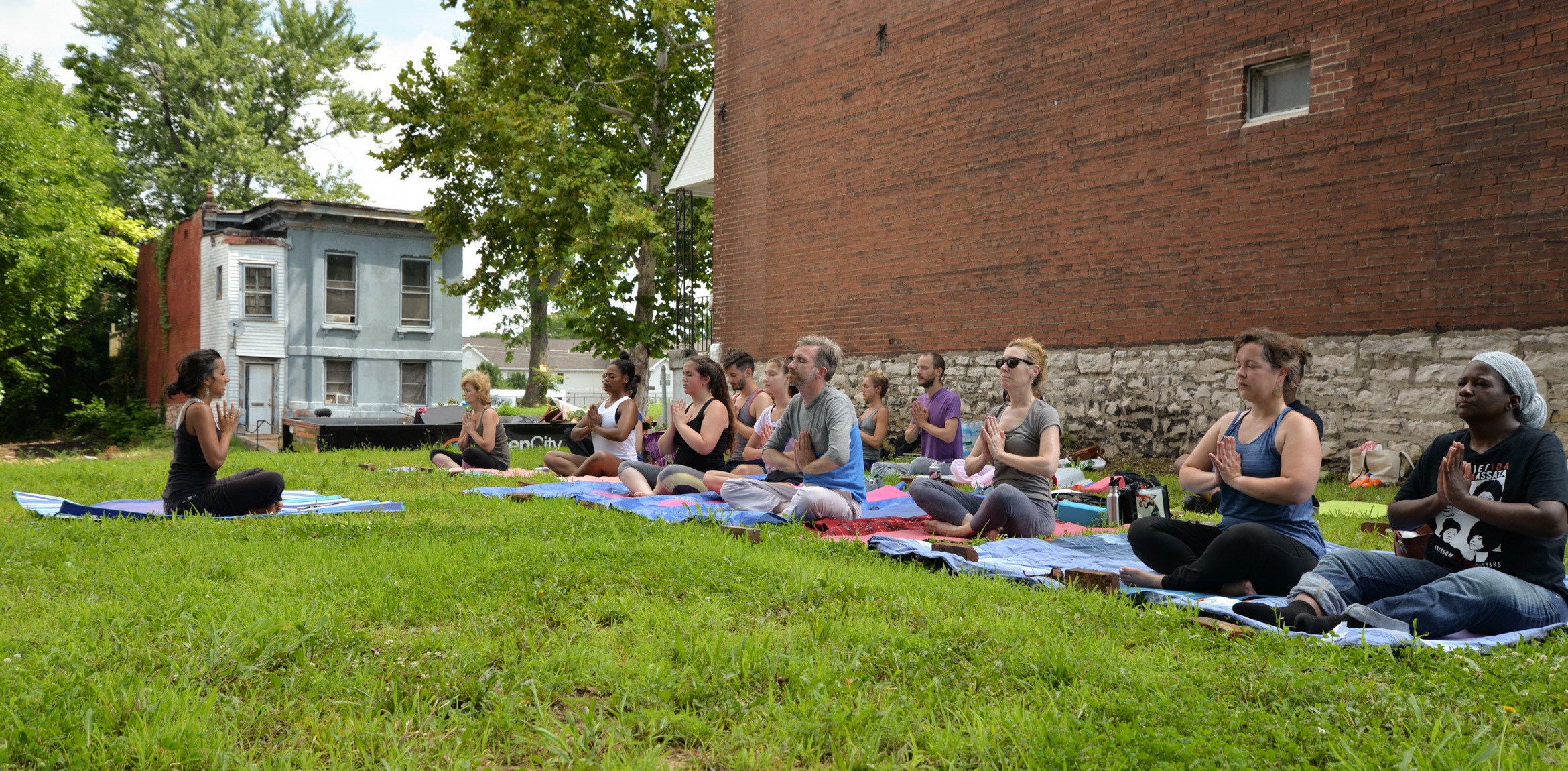 Mallory Nezam leads a yoga class on a lawn next to a brick building. The group sits cross-legged and holds their hands in a praying pose.
