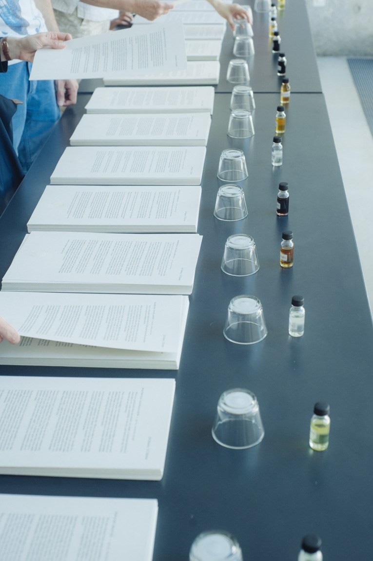 A closeup of small essential oil bottles and small clear plastic cups, and stacks of papers with correlated descriptions, sit on a long black table.