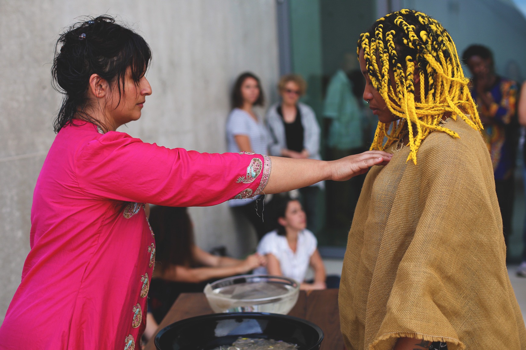 Bhanu Kapil performing a ritual with a participant, standing on either side of a table with bowls of water.