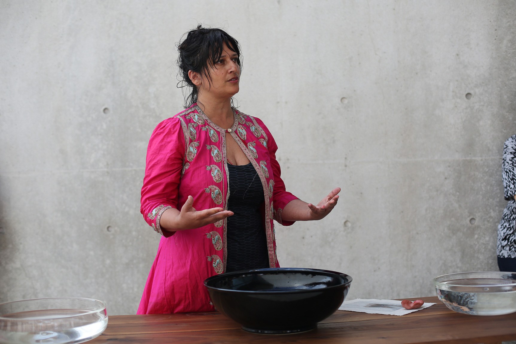 Bhanu Kapil stands behind a table with three bowls of water, speaking to the audience with her hands.