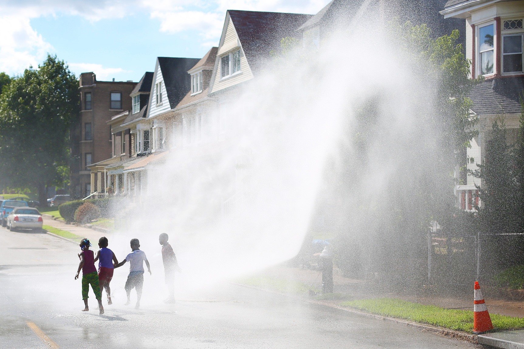 Children run through a large misty sprinkler in the street.