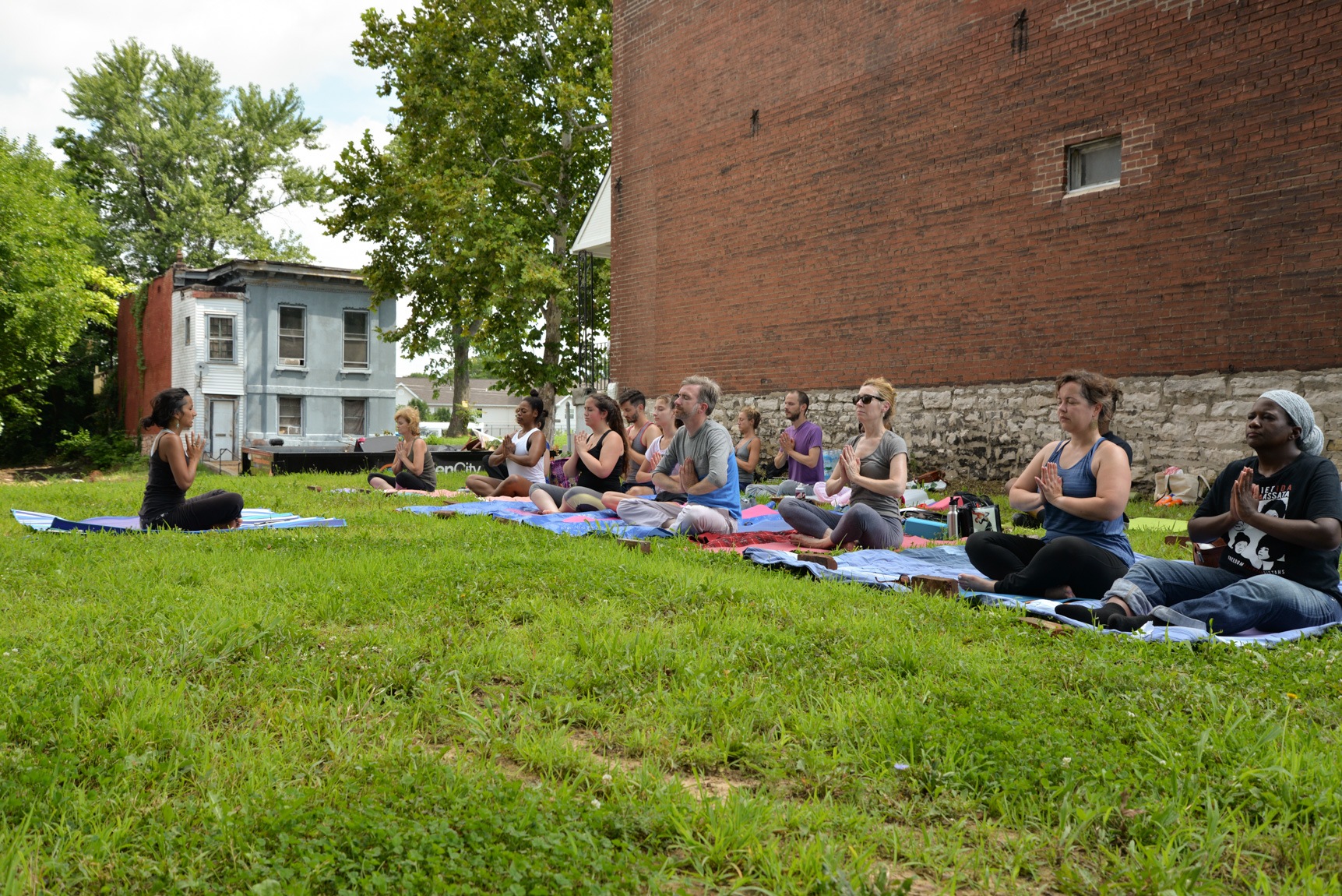 Mallory Nezam leads a gathering in a yoga exercise on a lawn in a residential neighborhood as participants sit cross-legged and their hands are in a praying pose.
