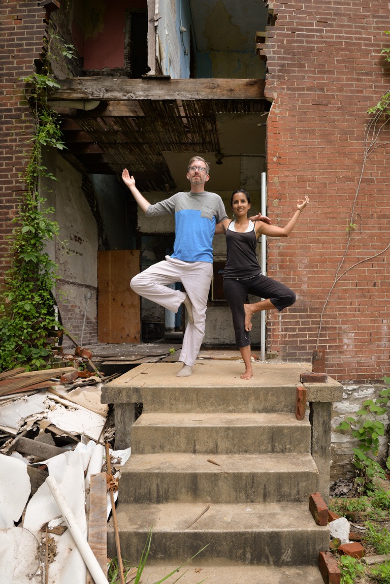 Mallory Nezam and Michael Allen pose for the camera in Tree Pose with their arms around each other's back on the steps of an abandoned building.