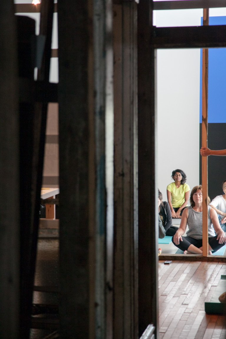The yoga participants sit on mats in the Main Gallery, framed by the installation " 4562 Enright Avenue."