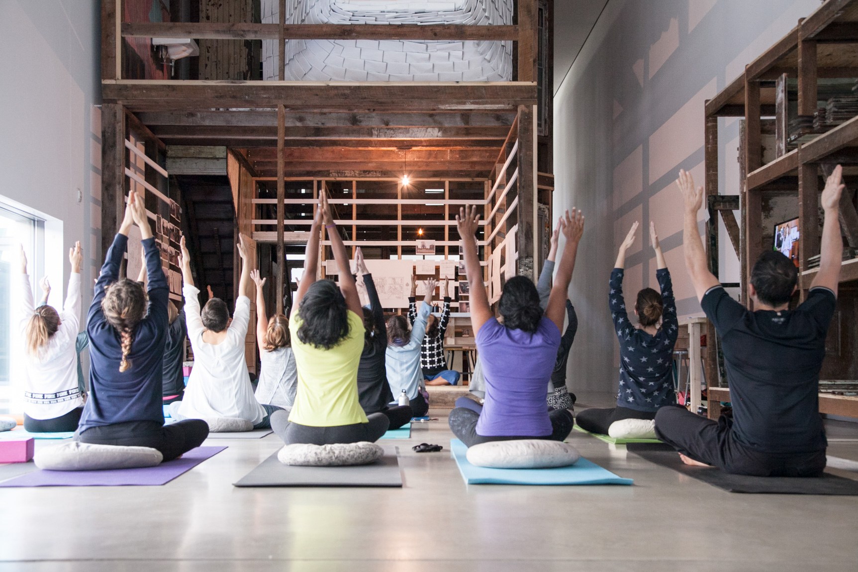 Mallory Nezam leads a group of participants in a yoga exercise in the Main Gallery, in front of the installation "4562 Enright Avenue." The group faces Nezam with their legs crossed and arms raised to the ceiling.