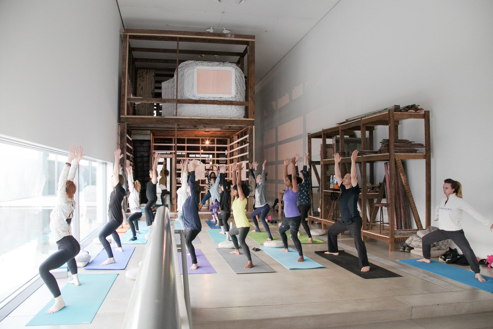 Attendants stand on yoga mats facing the windows adjoining the Water Court in the Main Gallery, their legs out and bent and arms straight up.