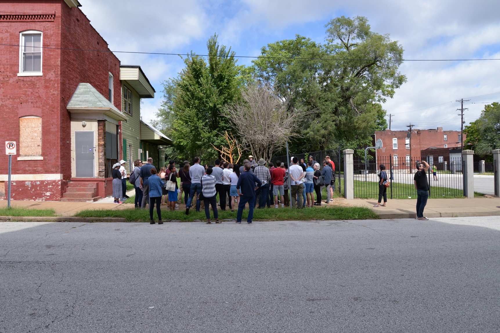 A large group gathers to listen to Amanda Williams and Andres L. Hernandez in a grassy area of a neighborhood.