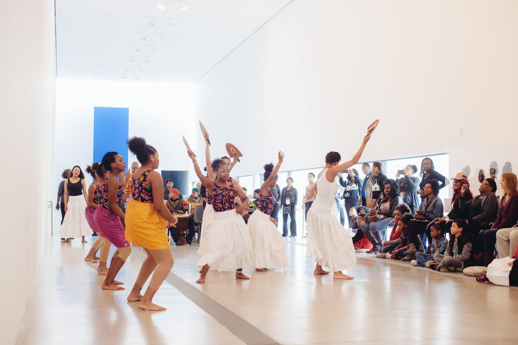 A large group of dancers perform an African inspired dance in the Main Gallery for an audience.