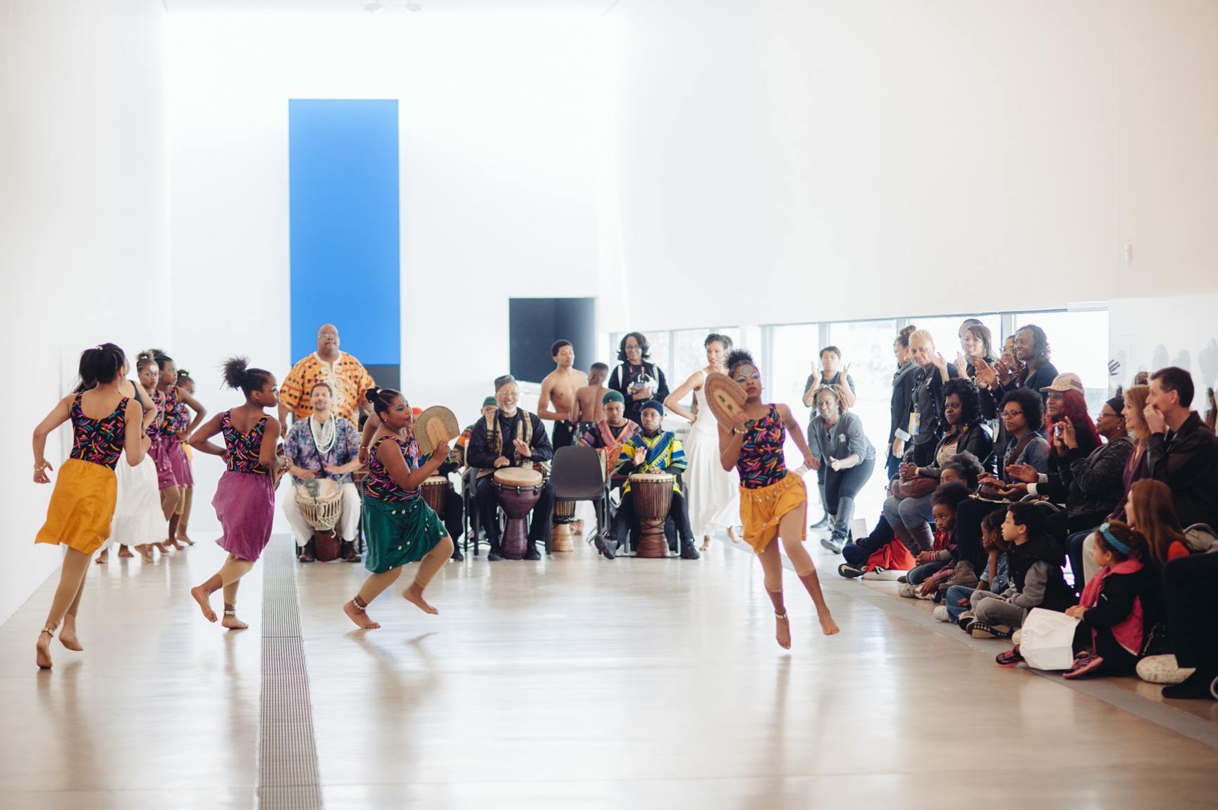A large group of dancers perform an African inspired dance in the Main Gallery for an audience.