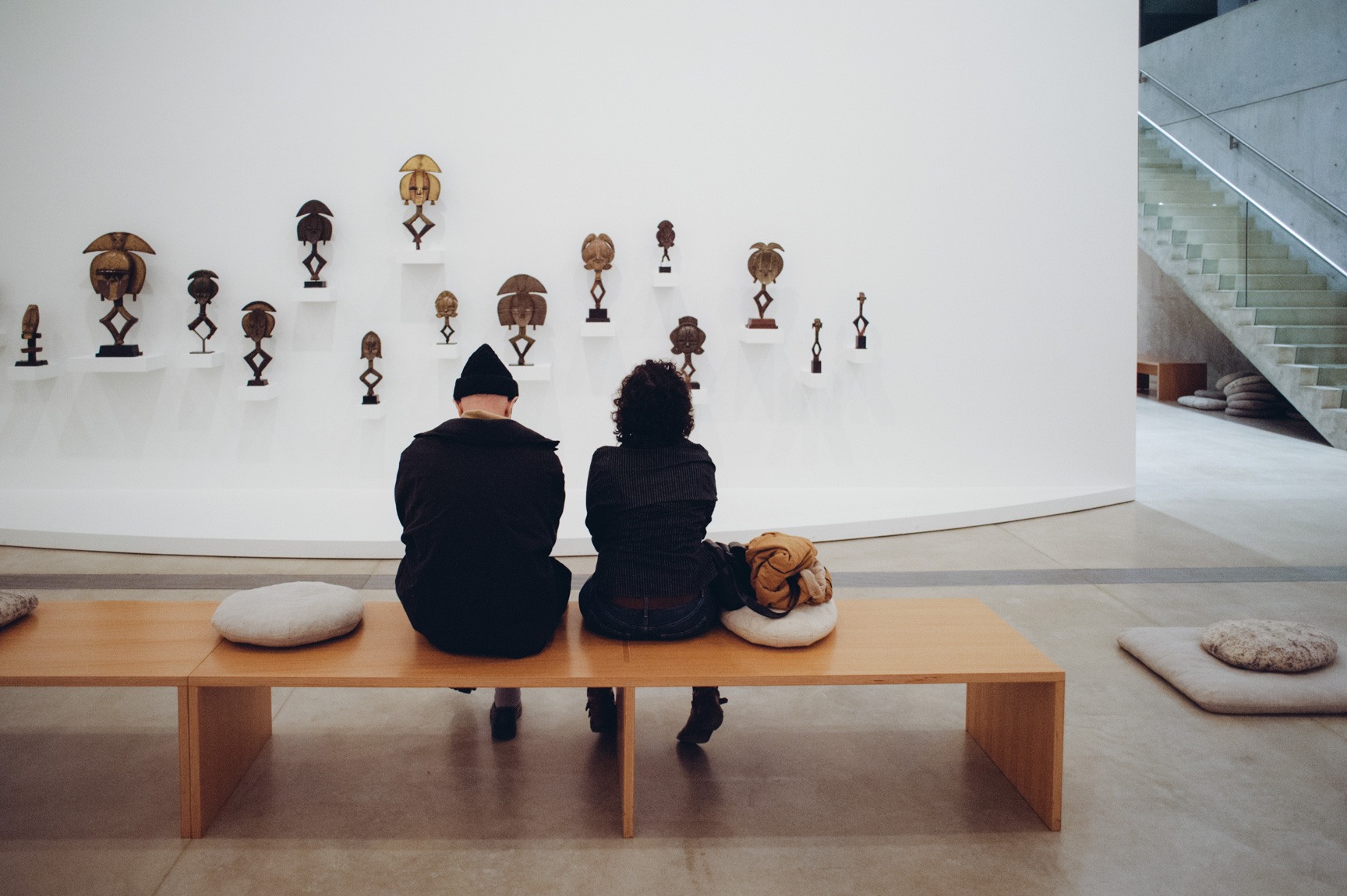 Two visitors sit on a wooden bench and view the wall of displayed African sculptures in the Main Gallery.