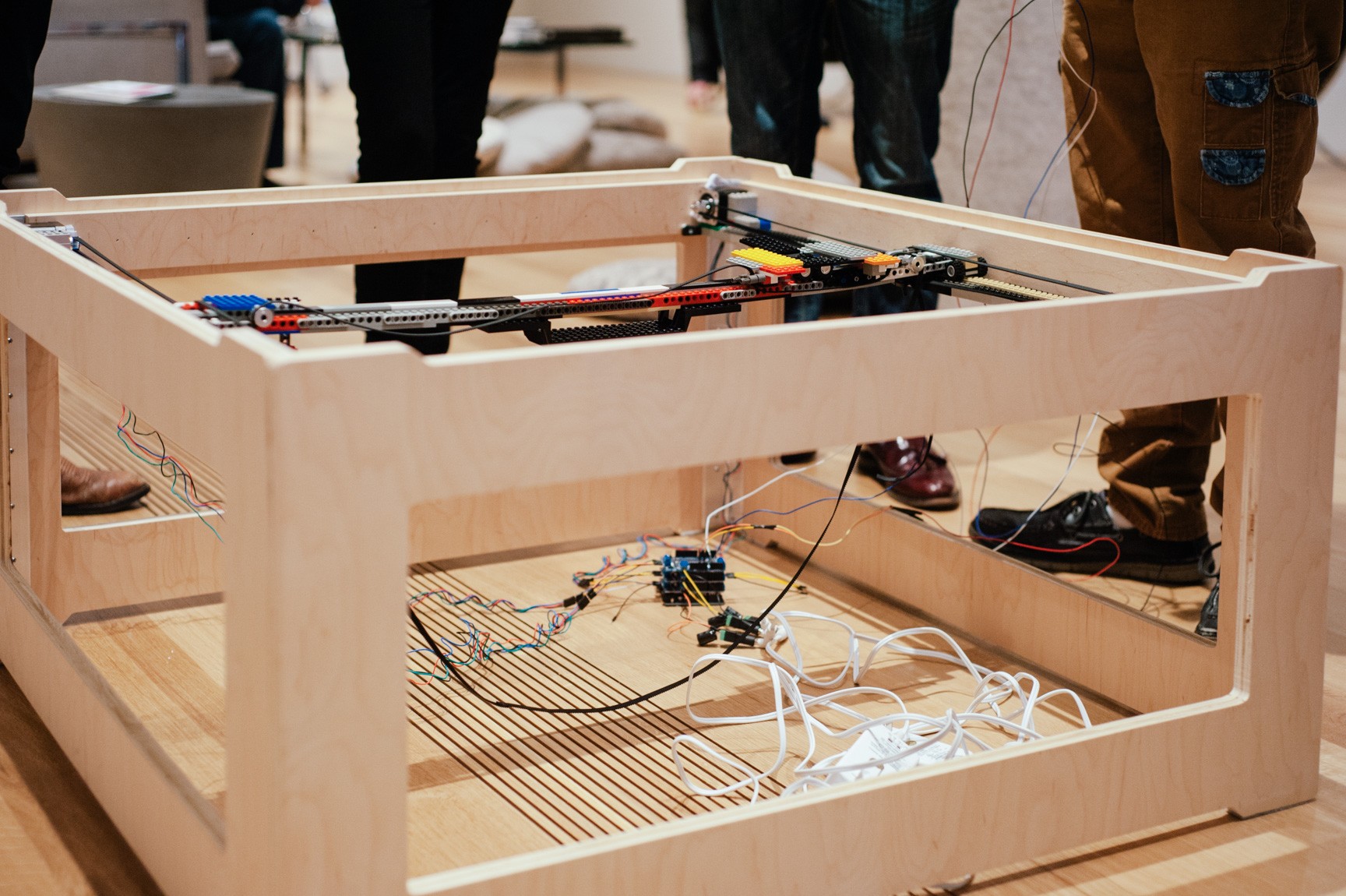 Collaborators stand around a homemade square wooden 3-D printer with wires hanging around it.