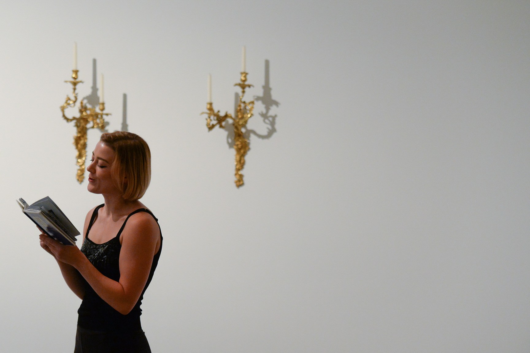 A presenter reads from "La Petite Maison" in front of two wall candelabra.