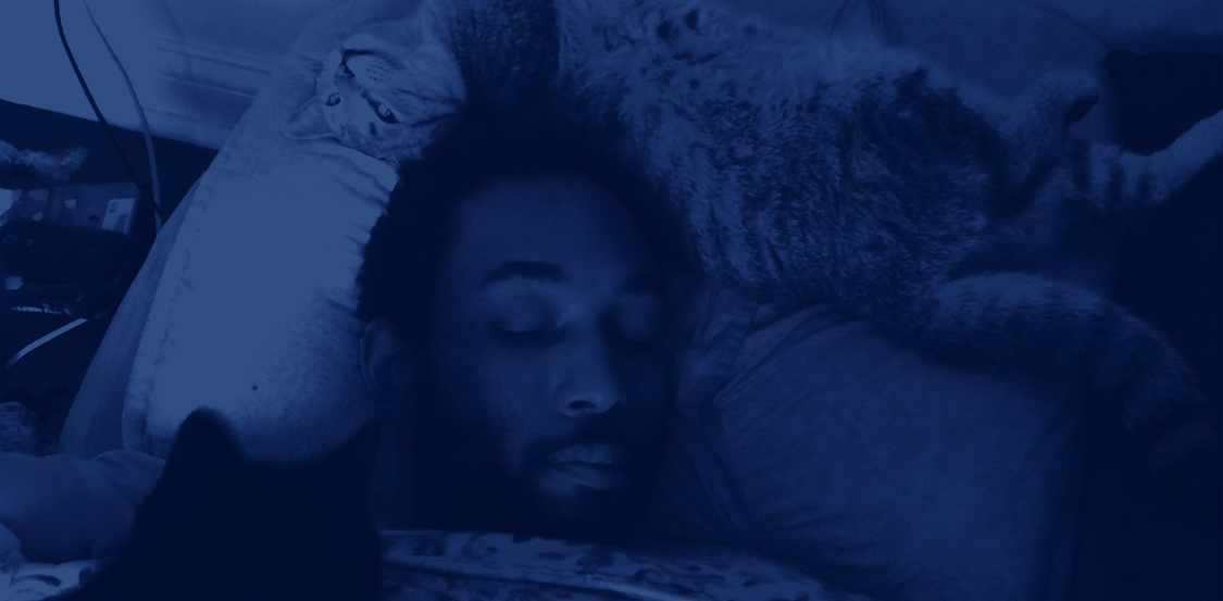 A blue photo of Frison in bed with a cat behind his head and a cat on his chest.