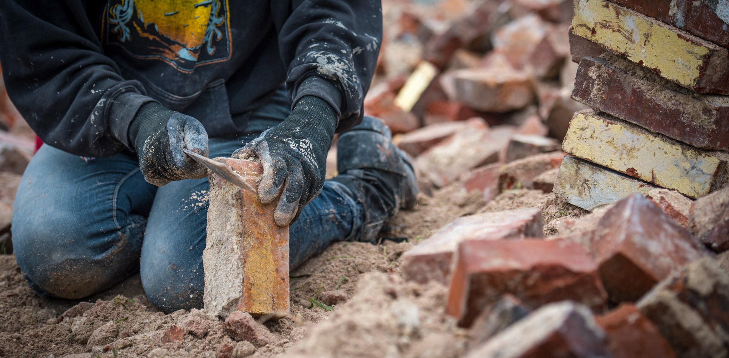A participant kneels in a pile of bricks and dirt, holding a brick in their hand and grinding a knife against it.