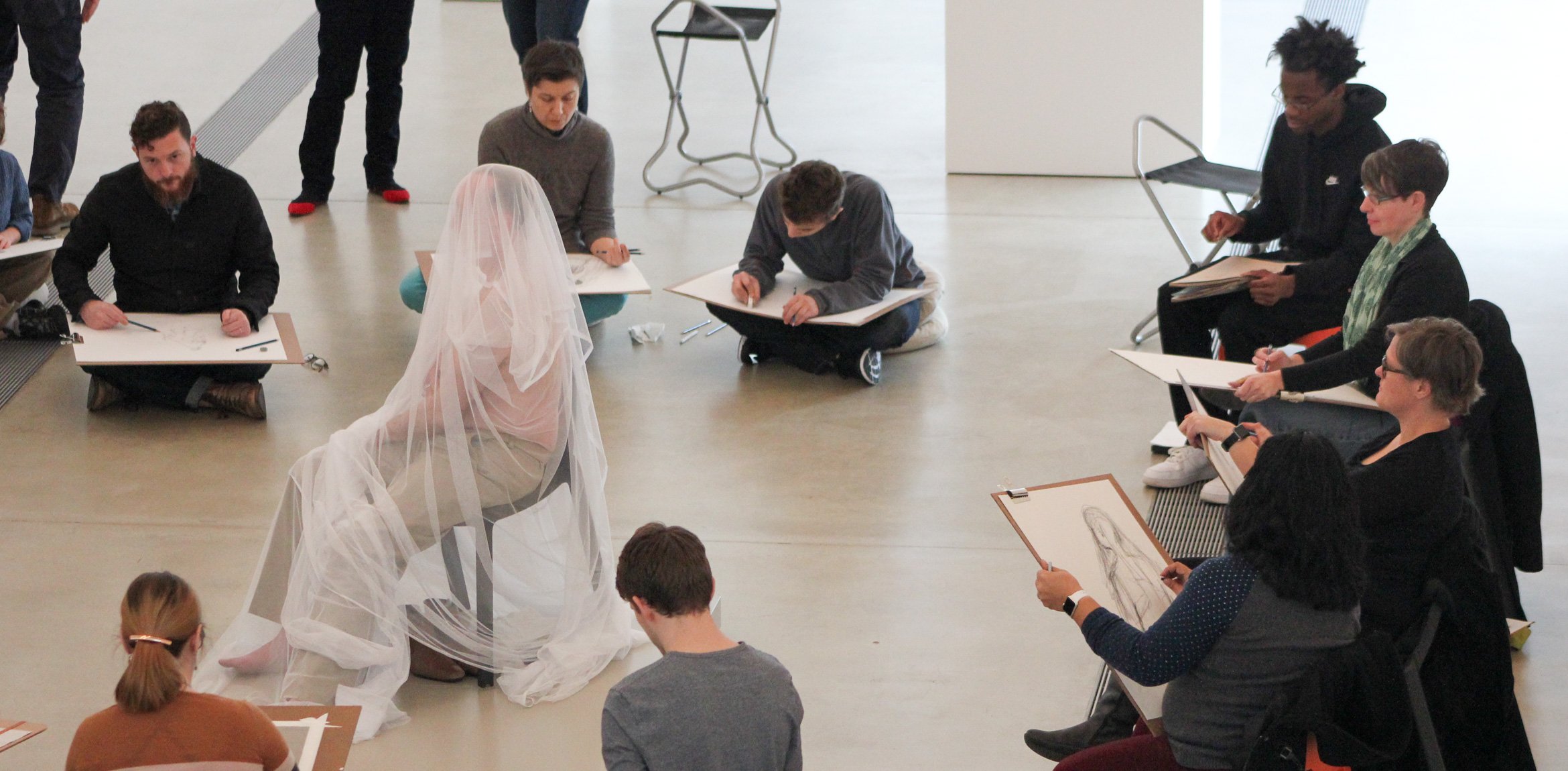 A circle of artists sketch a seated model, covered in sheer fabric in the Main Gallery.