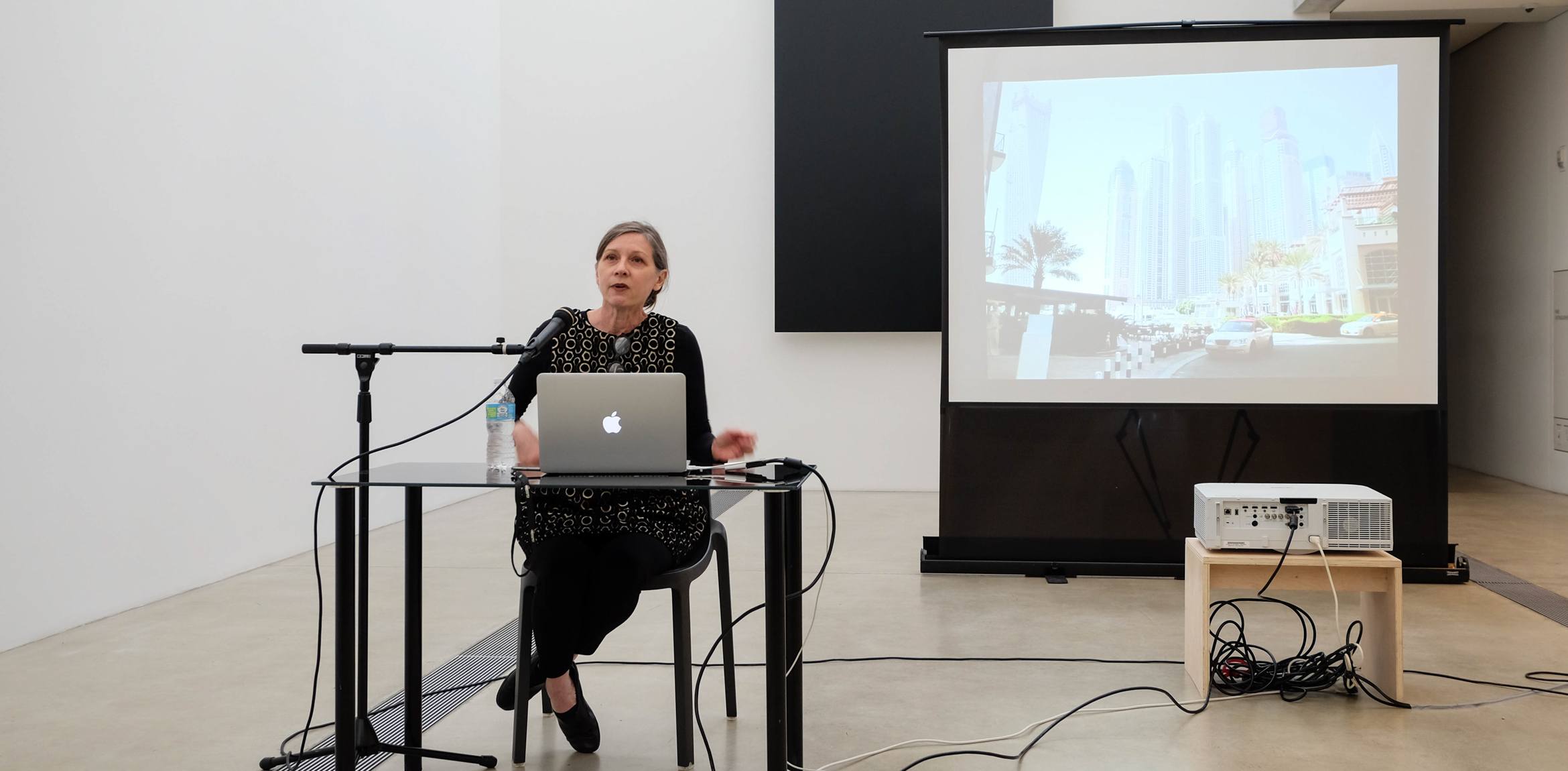 Keller Easterling sits at a small table with her laptop in front of Ellsworth Kelly's "Blue Black" and speaks into a microphone, a projector with supplemental images behind her.