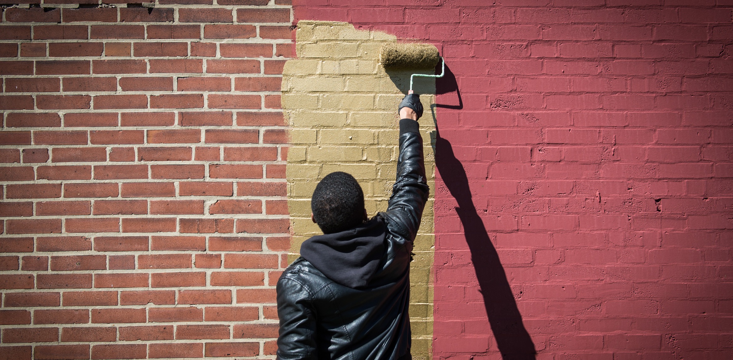 Amanda Williams uses a paint roller to paint a brick wall gold.