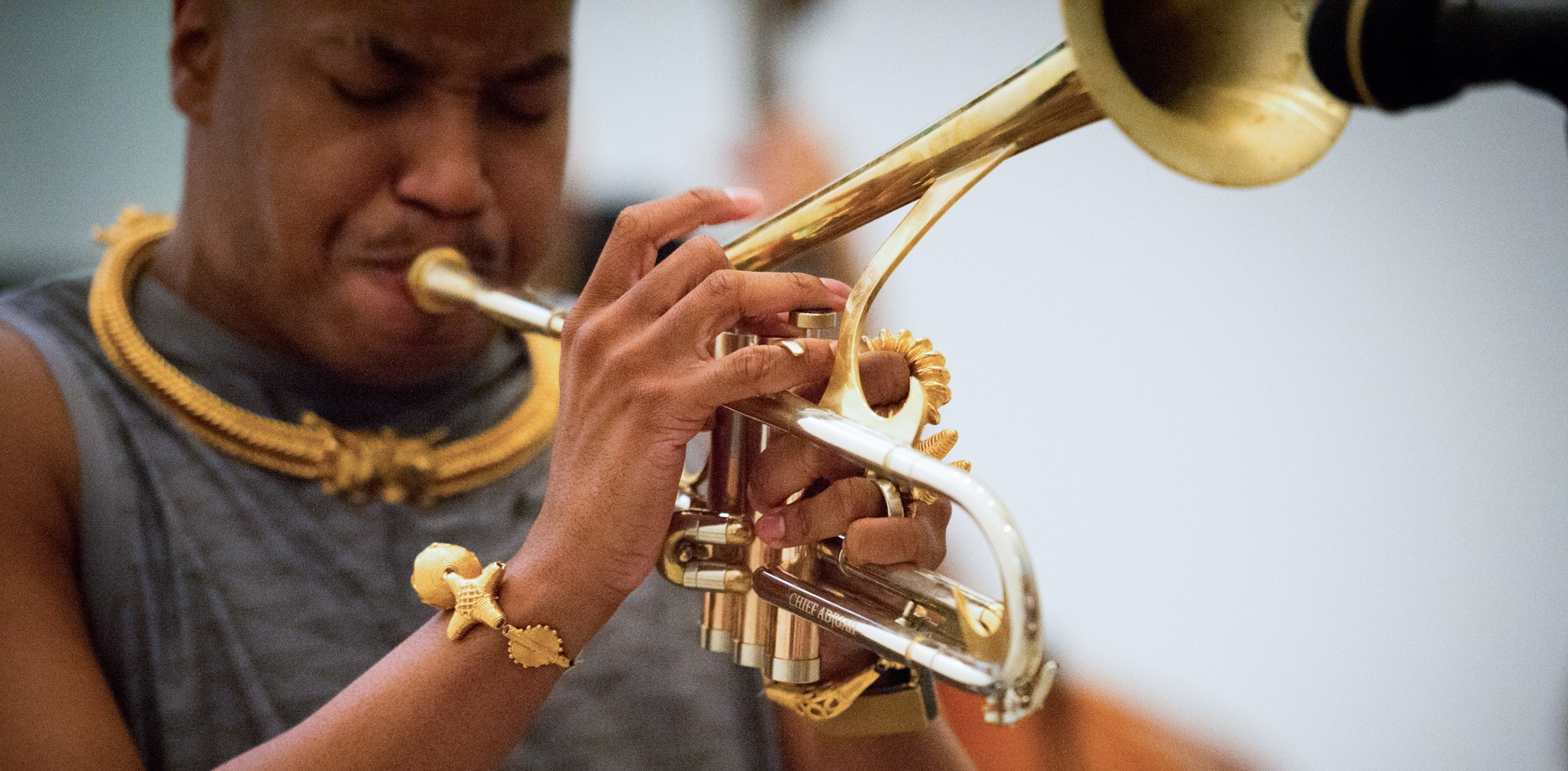 Christian Scott aTunde Adjuah plays the trumpet into a microphone. He wears a golden necklace, bracelets, and rings that match his trumpet.