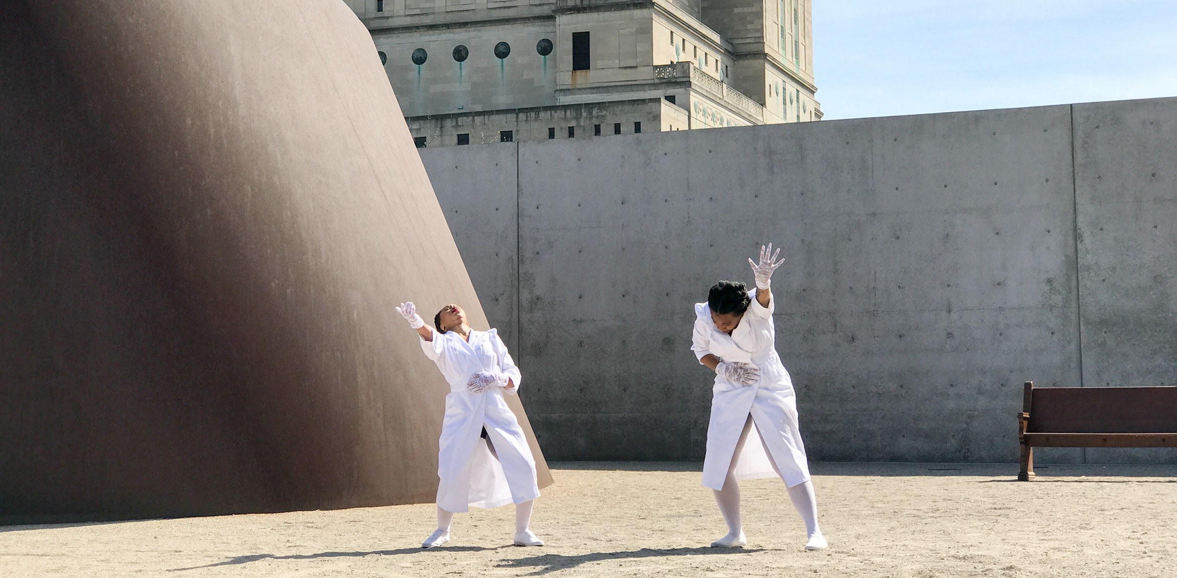 Two AUNTS dancers perform wearing white outfits and gloves beside Richard Serra's 
