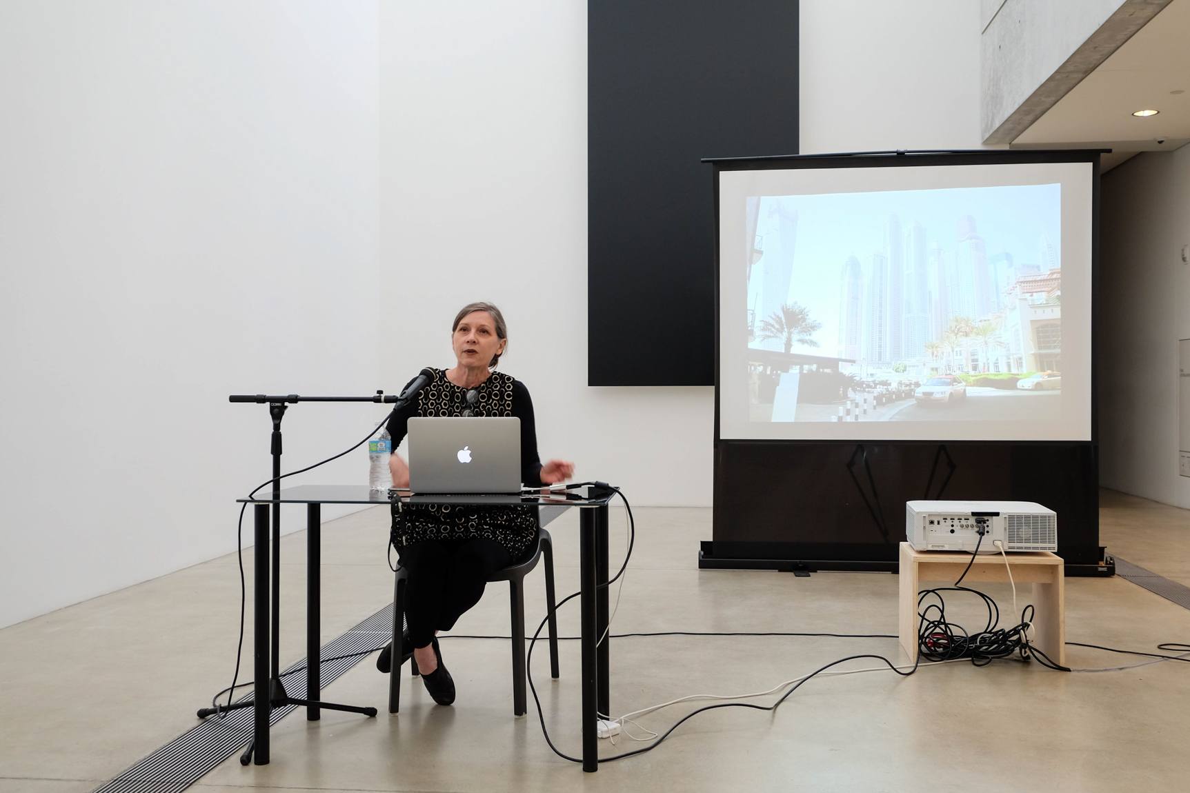 Keller Easterling sits in the Lower-Main Gallery at a small table in front of a projection screen and speaks into a microphone.