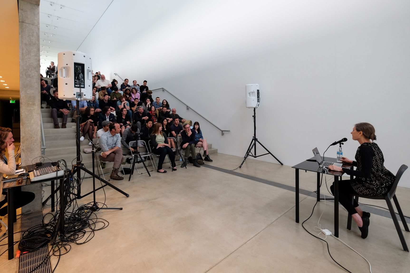 Keller Easterling gives a presentation in the Lower-Main Gallery to an audience on the Main Staircase. Easterling is seated behind a table with a microphone.