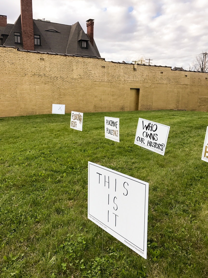 Five white placards sit in a lawn with a brick wall behind them.
