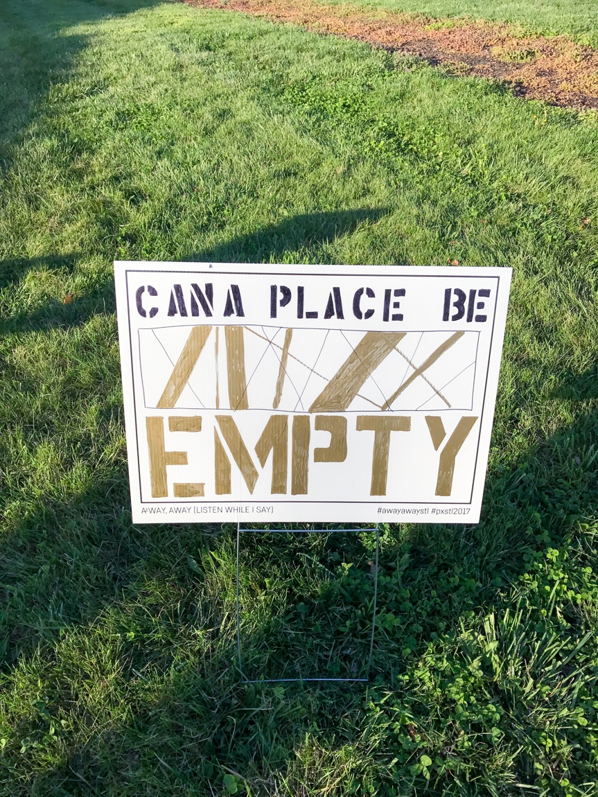 A white placard that reads "CAN A PLACE BE EMPTY" with a geometrical line design in a grass field.
