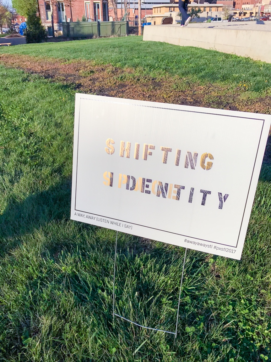 A white placard in a lawn that reads "SHIFTING IDENTITY." In the spaces between the first five letters of "identity," printed in a lighter color, it reads "SPACE."